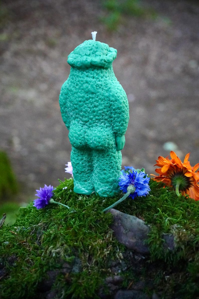 Op de afbeelding: Een groene kaars in de vorm van een persoon met een getextureerd oppervlak. De kaars staat op een bed van mos met een paar bloemen eromheen.