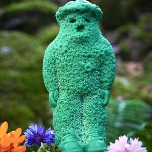 Op de afbeelding: Een groene kaars in de vorm van een persoon met een bloemenpatroon. De kaars staat op een bed van mos en bloemen.