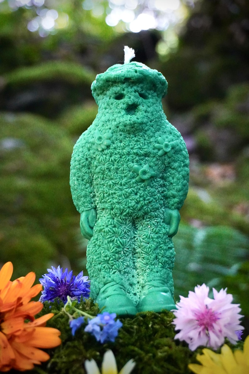 Op de afbeelding: Een groene kaars in de vorm van een persoon met een bloemenpatroon. De kaars staat op een bed van mos en bloemen.