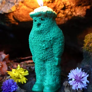Op de afbeelding: Een groene kaars in de vorm van een persoon met een vlam erop. De kaars is omringd door bloemen en bladeren.