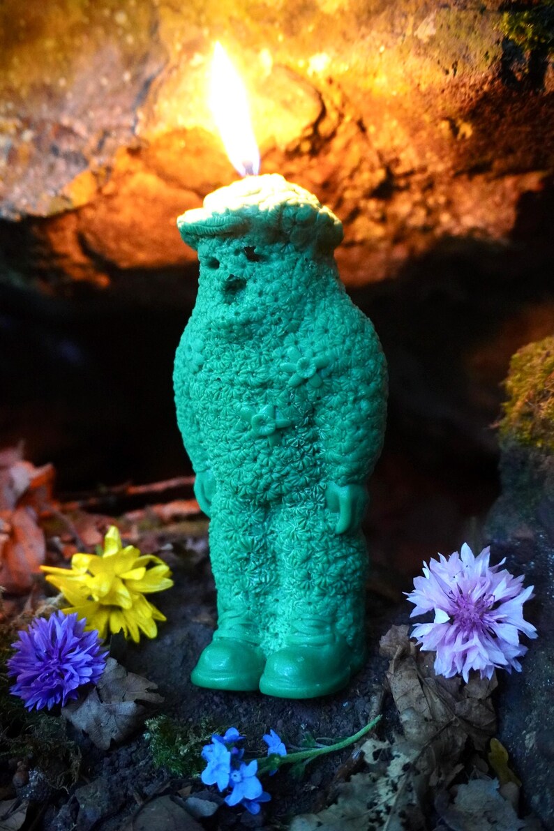Op de afbeelding: Een groene kaars in de vorm van een persoon met een vlam erop. De kaars is omringd door bloemen en bladeren.