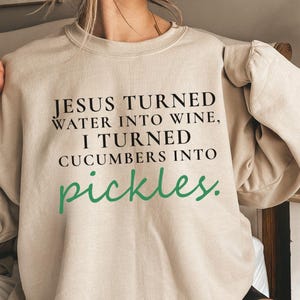May include: A beige sweatshirt featuring the text "JESUS TURNED WATER INTO WINE, I TURNED CUCUMBERS INTO pickles" in black and green. The sweatshirt has a crew neck and long sleeves.