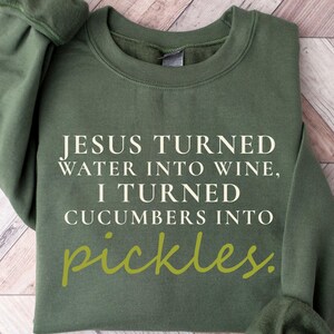 May include: An olive green sweatshirt with the text "JESUS TURNED WATER INTO WINE, I TURNED CUCUMBERS INTO pickles." The word "pickles" is in a lighter green script. The sweatshirt is folded on a light brown and white wooden surface.