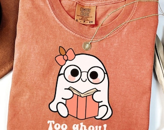 Puede incluir: Camiseta de color coral con un dibujo de un fantasma de dibujos animados con gafas y un lazo, leyendo un libro. El texto "Too ghoul for school" está impreso debajo. Un collar de oro está drapeado sobre la camiseta.