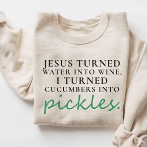 May include: A cream-coloured sweatshirt featuring the text "JESUS TURNED WATER INTO WINE, I TURNED CUCUMBERS INTO pickles." in black and green. The sweatshirt has a crew neck and long sleeves, with the left sleeve folded.