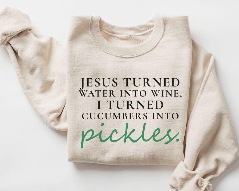 May include: A cream-coloured sweatshirt featuring the text "JESUS TURNED WATER INTO WINE, I TURNED CUCUMBERS INTO pickles." in black and green. The sweatshirt has a crew neck and long sleeves, with the left sleeve folded.