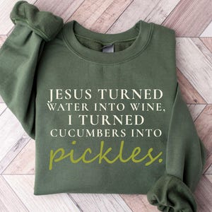 May include: An olive green sweatshirt with the text "JESUS TURNED WATER INTO WINE, I TURNED CUCUMBERS INTO pickles." The word "pickles" is in a lighter green script. The sweatshirt is folded on a light brown and white wooden surface.