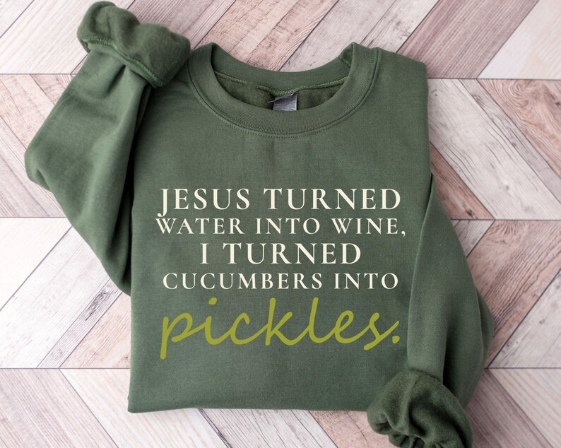 May include: An olive green sweatshirt with the text "JESUS TURNED WATER INTO WINE, I TURNED CUCUMBERS INTO pickles." The word "pickles" is in a lighter green script. The sweatshirt is folded on a light brown and white wooden surface.