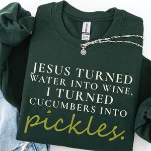 May include: A dark green sweatshirt with the text "JESUS TURNED WATER INTO WINE, I TURNED CUCUMBERS INTO pickles." A silver necklace with a sun pendant is draped across the sweatshirt. The sweatshirt is folded with a pair of blue jeans and another green sweatshirt.