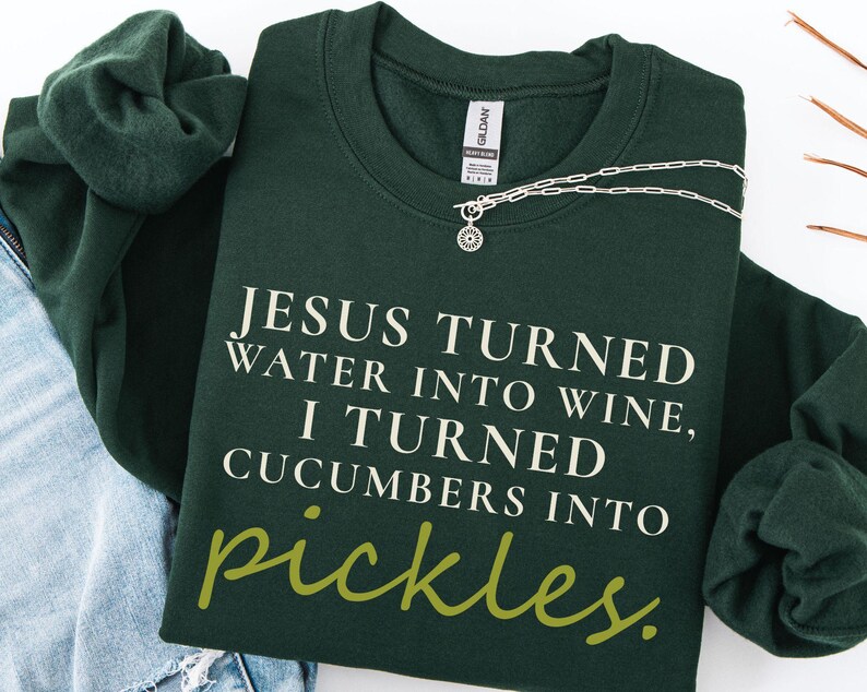 May include: A dark green sweatshirt with the text "JESUS TURNED WATER INTO WINE, I TURNED CUCUMBERS INTO pickles." A silver necklace with a sun pendant is draped across the sweatshirt. The sweatshirt is folded with a pair of blue jeans and another green sweatshirt.
