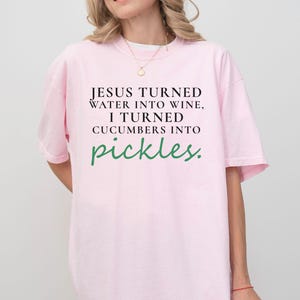 May include: A pale pink t-shirt featuring the quote "JESUS TURNED WATER INTO WINE, I TURNED CUCUMBERS INTO pickles." The text is in black and green. The shirt has a loose fit and short sleeves.