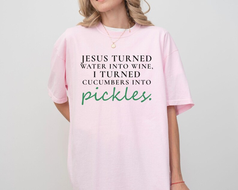 May include: A pale pink t-shirt featuring the quote "JESUS TURNED WATER INTO WINE, I TURNED CUCUMBERS INTO pickles." The text is in black and green. The shirt has a loose fit and short sleeves.