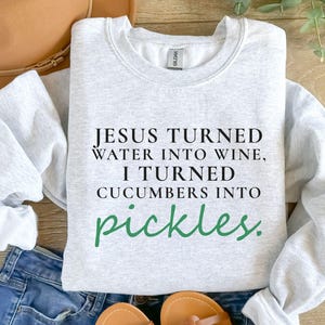 May include: Light grey sweatshirt featuring the text "JESUS TURNED WATER INTO WINE, I TURNED CUCUMBERS INTO pickles." in black and green. The sweatshirt is folded on blue jeans and a brown hat.