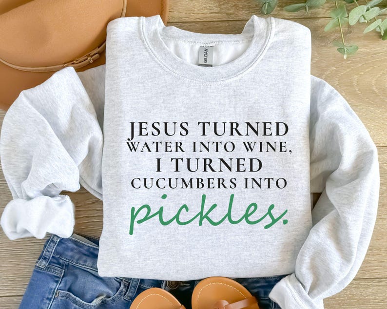 May include: Light grey sweatshirt featuring the text "JESUS TURNED WATER INTO WINE, I TURNED CUCUMBERS INTO pickles." in black and green. The sweatshirt is folded on blue jeans and a brown hat.