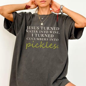 May include: Dark grey oversized t-shirt with the text "JESUS TURNED WATER INTO WINE, I TURNED CUCUMBERS INTO pickles." The word "pickles" is in a green, handwritten font. The shirt has short sleeves and a relaxed fit.