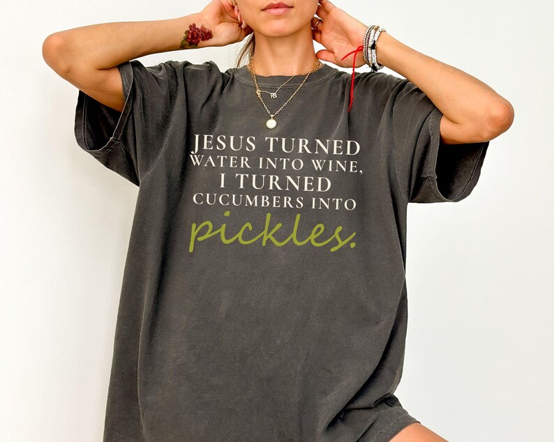 May include: Dark grey oversized t-shirt with the text "JESUS TURNED WATER INTO WINE, I TURNED CUCUMBERS INTO pickles." The word "pickles" is in a green, handwritten font. The shirt has short sleeves and a relaxed fit.