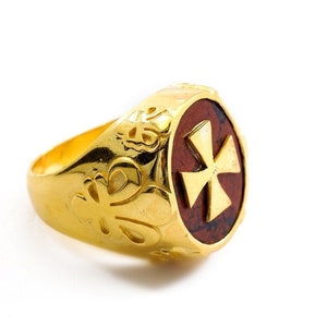 Könnte beinhalten: Ein goldener Ring mit einem roten Stein, der in der Mitte eingesetzt ist. Der Stein hat ein goldenes Kreuzdesign. Die Ringschiene ist mit komplizierten Blumenmustern verziert.