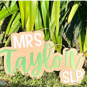 May include: Two wooden signs with white text. The larger sign says "Mrs. Taylor" in green cursive lettering. The smaller sign says "SLP" in white lettering.
