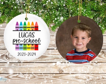 May include: White ceramic ornament with a rainbow of crayons and the text "Lucas pre-school 2023-2024". A second ornament features a photo of a young boy.
