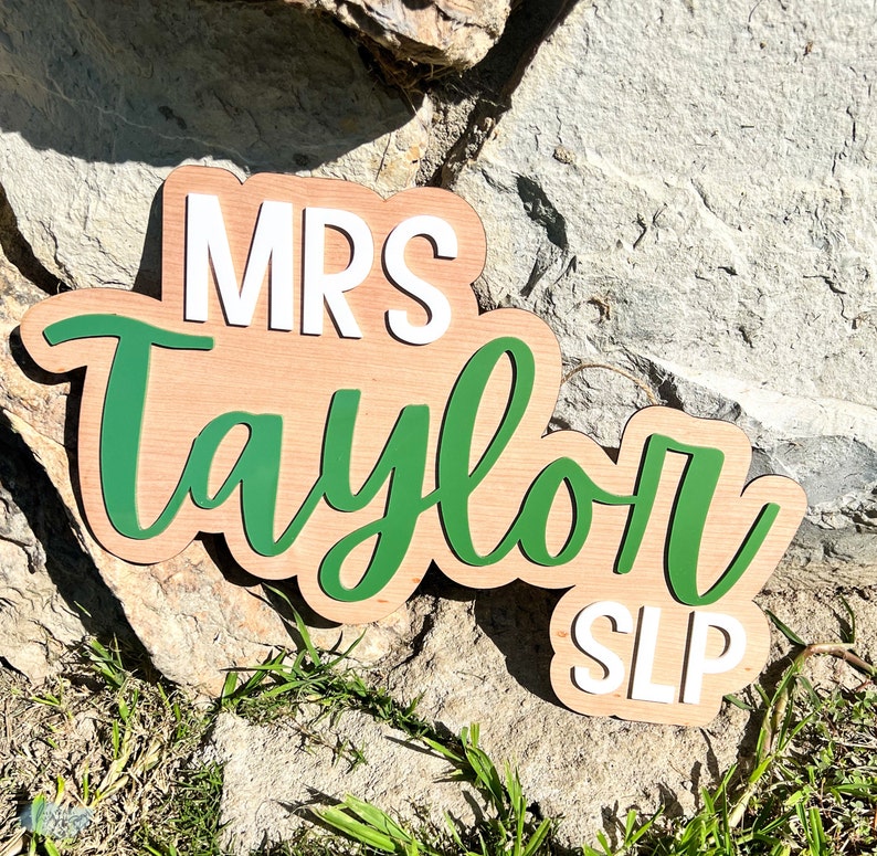 May include: Wooden sign with the text "MRS Taylor SLP" in white and green lettering on a natural wood background.