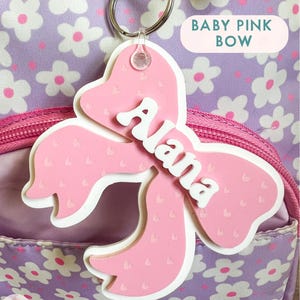 May include: A pink bow-shaped name tag with the name "Alana" written in white letters. The tag is attached to a silver key ring and has a small clear plastic loop.