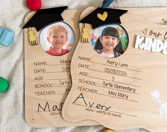 May include: Wooden kindergarten photo frames with a graduation cap design. Each frame has a space for a photo, name, date, age, school, and teacher. One frame has the name "Mason", the other "Avery Lynn".