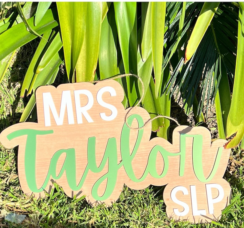 May include: Two wooden signs with white text. The larger sign says "Mrs. Taylor" in green cursive lettering. The smaller sign says "SLP" in white lettering.