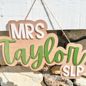 May include: A wooden sign with the text "MRS Taylor SLP" in white letters on a green background. The sign is hanging from a string.