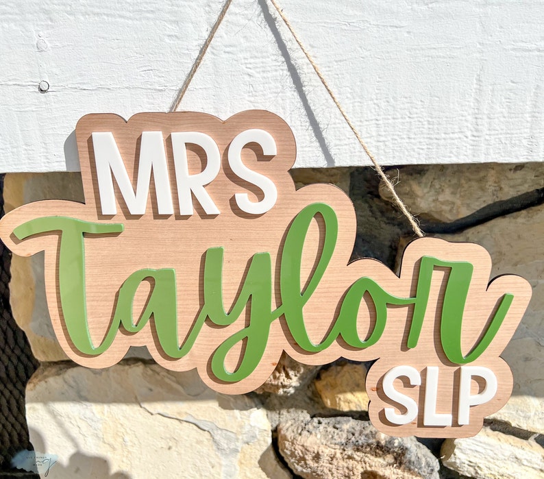 May include: A wooden sign with the text "MRS Taylor SLP" in white letters on a green background. The sign is hanging from a string.