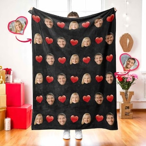 Puede incluir: Manta negra con un patrón repetido de corazones rojos y caras. Las caras son de un hombre y una mujer. La manta está sujeta por una persona. Un regalo personalizado.