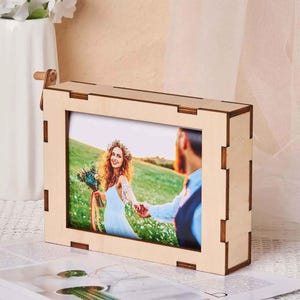 May include: A wooden photo frame with a built-in music box mechanism. The frame holds a photo of a couple in a field. The frame is light brown and has a simple design.