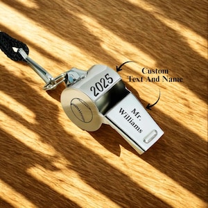 May include: A silver baseball whistle with a black lanyard. The whistle has a baseball design on the side and the text "2025" on top. The text "Mr. Williams" is printed on the front. The image is on a wooden surface.