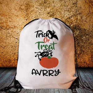 Peut inclure: Sac blanc à cordon avec un thème d'Halloween. Le sac présente les mots "Trick or Treat" en noir, vert et rouge, ainsi qu'un dessin de chauve-souris, d'araignée et de citrouille. Le nom "AVRRY" est imprimé en bas.