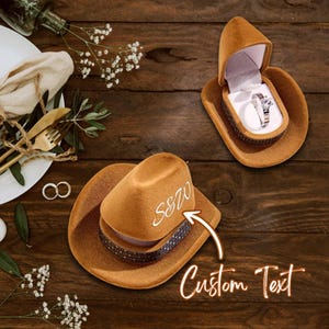 Puede incluir: Una caja de anillos con forma de sombrero de vaquero marrón con un anillo plateado en su interior. El sombrero tiene una banda decorativa y el texto "S&80" en la corona. También se ven las palabras "Custom Text". La caja de anillos está abierta, revelando un anillo de diamantes.