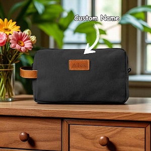 May include: Black toiletry bag with a brown leather handle and a small leather patch with the name "Alex". The bag has a rectangular shape and a zipper closure. The text "Custom Name" is written above the bag.