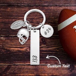 May include: Silver football-themed keychain with a football, helmet, and tag that says "Eli". A round tag has the number "34". A brown and white football is in the background.