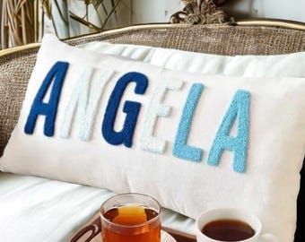 Könnte beinhalten: Ein weißes rechteckiges Kissen mit dem Namen "ANGELA" in strukturierten blauen und weißen Buchstaben gestickt. Ein Tablett mit einem Glas Tee und einer Teetasse befindet sich in der Nähe. Das Kissen liegt auf einem Bett.