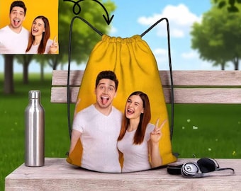 May include: Yellow drawstring backpack featuring a photo of a smiling couple. The bag has black drawstrings and a yellow background. A stainless steel water bottle and headphones are next to the bag on a wooden bench.
