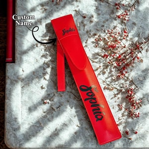 Puede incluir: Un estuche para bolígrafo de cuero rojo con el nombre "Sophia" impreso en escritura negra. El estuche tiene una correa larga adjunta y está sobre una superficie clara y texturizada. Bayas y ramas rojas decoran el fondo.