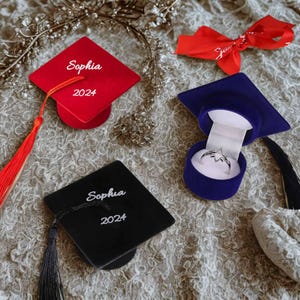 Könnte beinhalten: Ein Bild zum Thema Abschlussfeier mit einem roten und schwarzen Mortarboard, jeweils mit der Aufschrift "Sophia 2024". Ein blauer Mortarboard enthält eine Ringschatulle mit einem Silberring. Ein rotes Band sorgt für einen festlichen Touch.