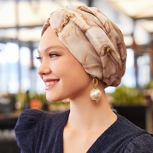 May include: A turban-style head covering in beige with gold accents. The fabric has a marbled pattern. The person is wearing gold hoop earrings with large, round, pearl-like pendants and a dark blue top.