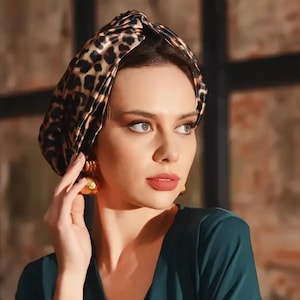 Könnte beinhalten: Eine Frau trägt ein dunkelblaugrünes Kleid und goldene Ohrringe. Sie trägt auch einen Turban mit Leopardenmuster. Das Kleid hat einen V-Ausschnitt und lange Ärmel. Die Frau schaut zur Seite.