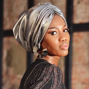 May include: A woman wearing a silver metallic turban and large silver hoop earrings. She is wearing a black top with a shimmering, striped pattern. The image is a portrait with a soft focus.