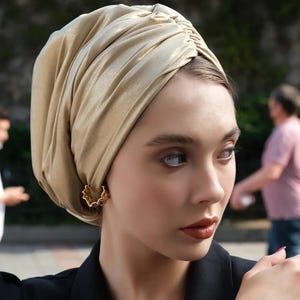 May include: A woman wearing a gold-colored turban and a black blazer. The turban is made of a soft, draped fabric. She is also wearing gold-colored earrings. The background is blurred, with other people visible.