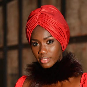 May include: A woman wearing a red turban and a red dress with a black fur collar. The turban is made of a shiny, textured fabric. The woman has dark skin and is looking at the camera.