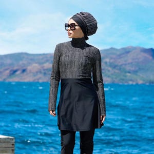 May include: A modest swimwear set in black and silver, featuring a long-sleeved top with vertical stripes, a black skirt, and leggings. The outfit includes a matching head covering and sunglasses. The person is standing on a wooden dock with a blue ocean and mountains in the background.