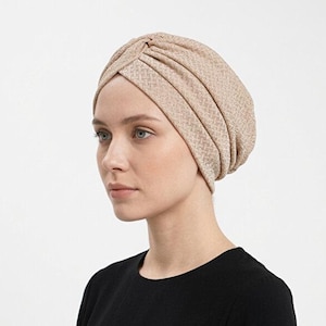 May include: A beige turban-style head covering with a textured pattern. The head wrap is made of a soft material and is worn by a person wearing a black top. The turban is a stylish accessory.