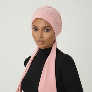 May include: A light pink head covering with a quilted design, paired with a matching scarf. The head covering is worn with a black long-sleeved top. The scarf drapes down the front.
