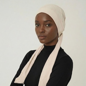 May include: A beige head covering with a quilted design on the crown and long, flowing ties. The head covering is paired with a black long-sleeved top. The overall aesthetic is modest and stylish.
