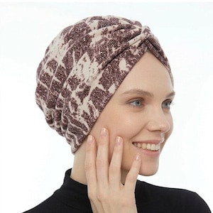 May include: A patterned turban in shades of burgundy and beige. The turban is made of a soft fabric and has a knotted design at the top. The person in the image is wearing a black turtleneck.